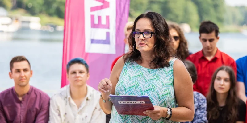Kobieta w okularach przemawia do zgromadzonych na brzegu jeziora, trzymając ulotkę z napisem Lewica, a w tle widać różowy baner i innych uczestników.