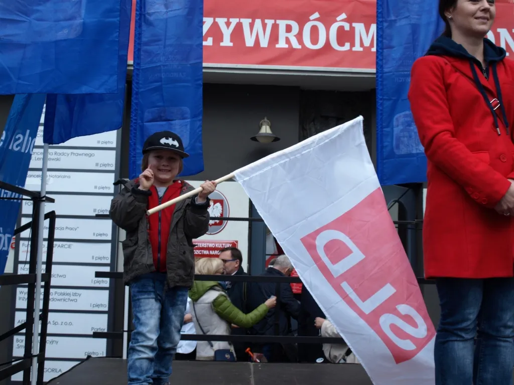 Uśmiechnięte dziecko w czapce stoi na scenie podczas obchodów Święta Pracy (1 maja 2017), trzymając biało‑czerwoną flagę z czerwonym napisem, obok stoi dorosła osoba w czerwonym płaszczu.