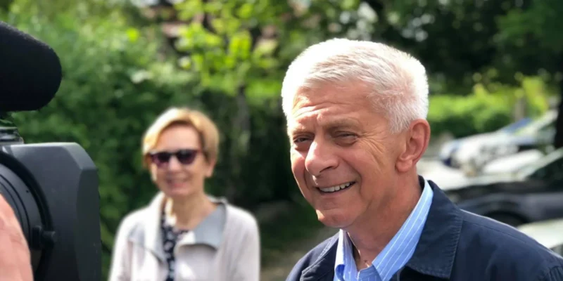 Ujęcie na zewnątrz: uśmiechnięty starszy mężczyzna w błękitnej koszuli i granatowej marynarce stoi obok kamery, a w tle widoczna jest kobieta w okularach przeciwsłonecznych.