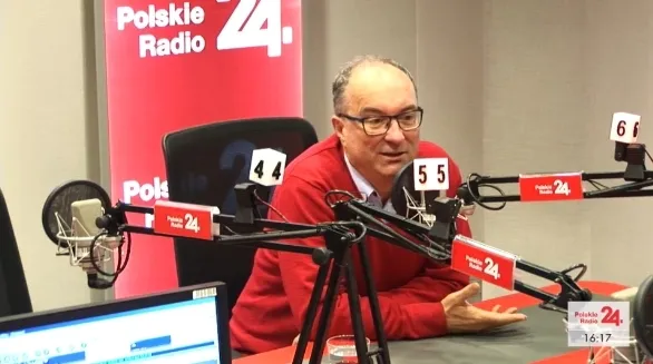 Mężczyzna w czerwonym swetrze gestykuluje i rozmawia do mikrofonu podczas nagrania w studiu Polskie Radio 24.