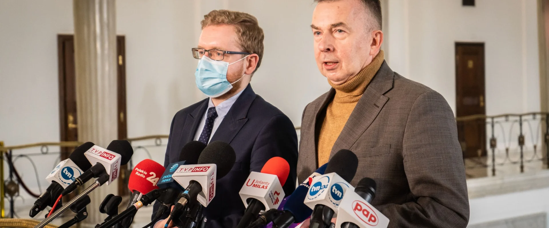 Dwaj mężczyźni w garniturach stoją przed mikrofonami podczas konferencji prasowej w eleganckim wnętrzu.