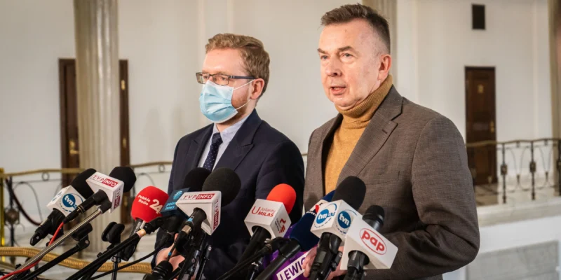 Dwaj mężczyźni w garniturach stoją przed mikrofonami podczas konferencji prasowej w eleganckim wnętrzu.