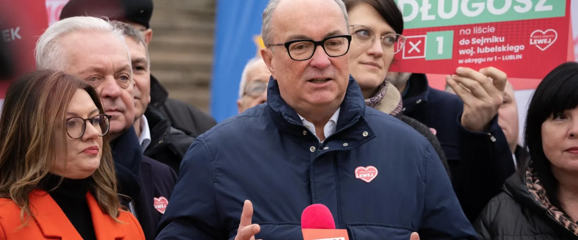 Mężczyzna w granatowej kurtce i okularach wygłasza przemówienie do mikrofonu podczas konferencji Lewicy, otoczony innymi politykami i plakatami związanymi z NATO.