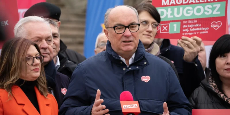 Mężczyzna w granatowej kurtce i okularach wygłasza przemówienie do mikrofonu podczas konferencji Lewicy, otoczony innymi politykami i plakatami związanymi z NATO.