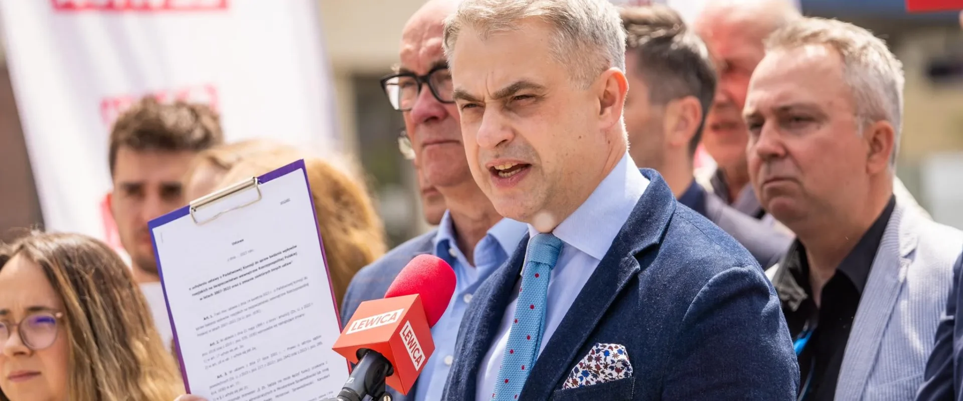 Mężczyzna w granatowym garniturze przemawia do mikrofonów na konferencji prasowej Lewicy, trzymając kartę z dokumentem, a w tle stoją inni uczestnicy i banery z napisem Lewica.