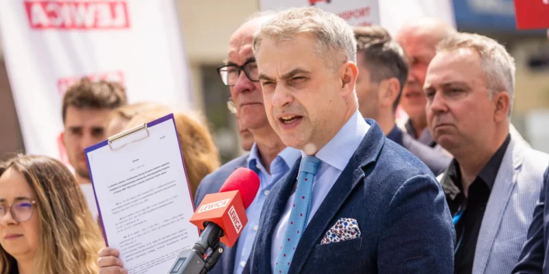 Mężczyzna w granatowym garniturze przemawia do mikrofonów na konferencji prasowej Lewicy, trzymając kartę z dokumentem, a w tle stoją inni uczestnicy i banery z napisem Lewica.