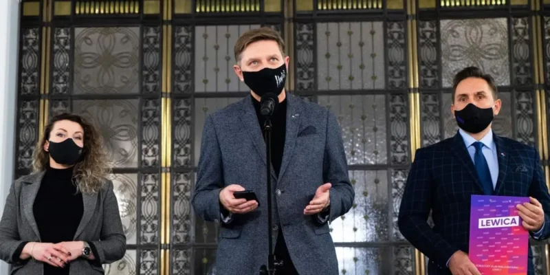 Trzy osoby w maseczkach stoją na konferencji: pośrodku mężczyzna przemawia do mikrofonu i trzyma telefon, z lewej kobieta, z prawej mężczyzna trzyma plik z napisem LEWICA.