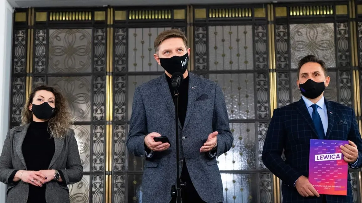 Trzy osoby w maseczkach stoją na konferencji: pośrodku mężczyzna przemawia do mikrofonu i trzyma telefon, z lewej kobieta, z prawej mężczyzna trzyma plik z napisem LEWICA.