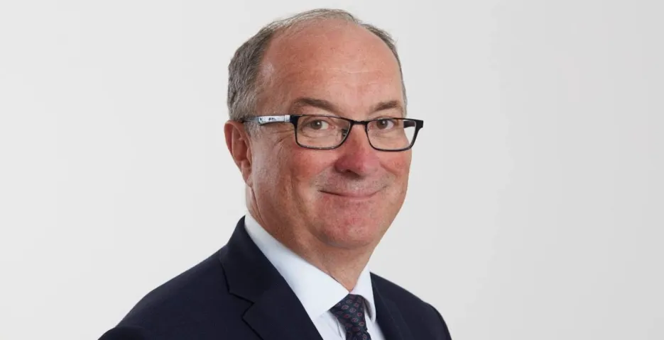 Portret mężczyzny w garniturze i okularach, uśmiechniętego na neutralnym białym tle.