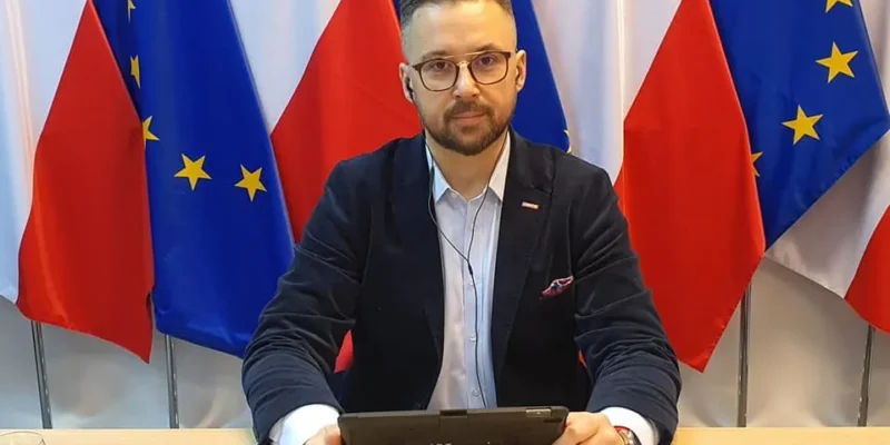 Mężczyzna w garniturze i okularach siedzi przy stole z tabletem; na tle widoczne są flagi Polski i Unii Europejskiej.