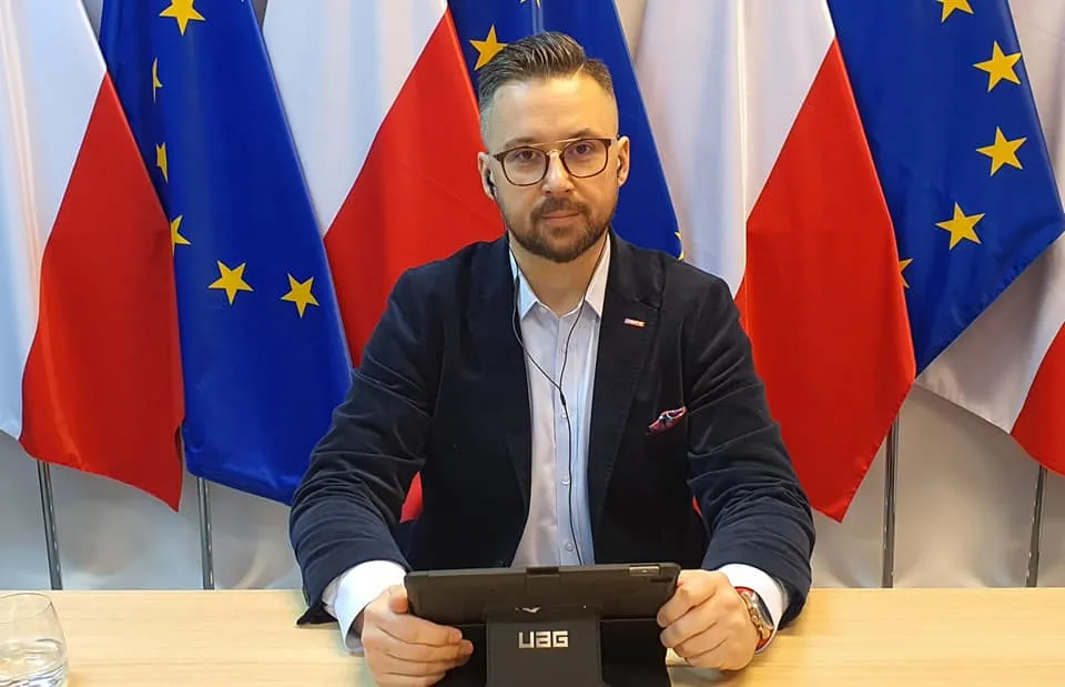 Mężczyzna w garniturze i okularach siedzi przy stole z tabletem; na tle widoczne są flagi Polski i Unii Europejskiej.