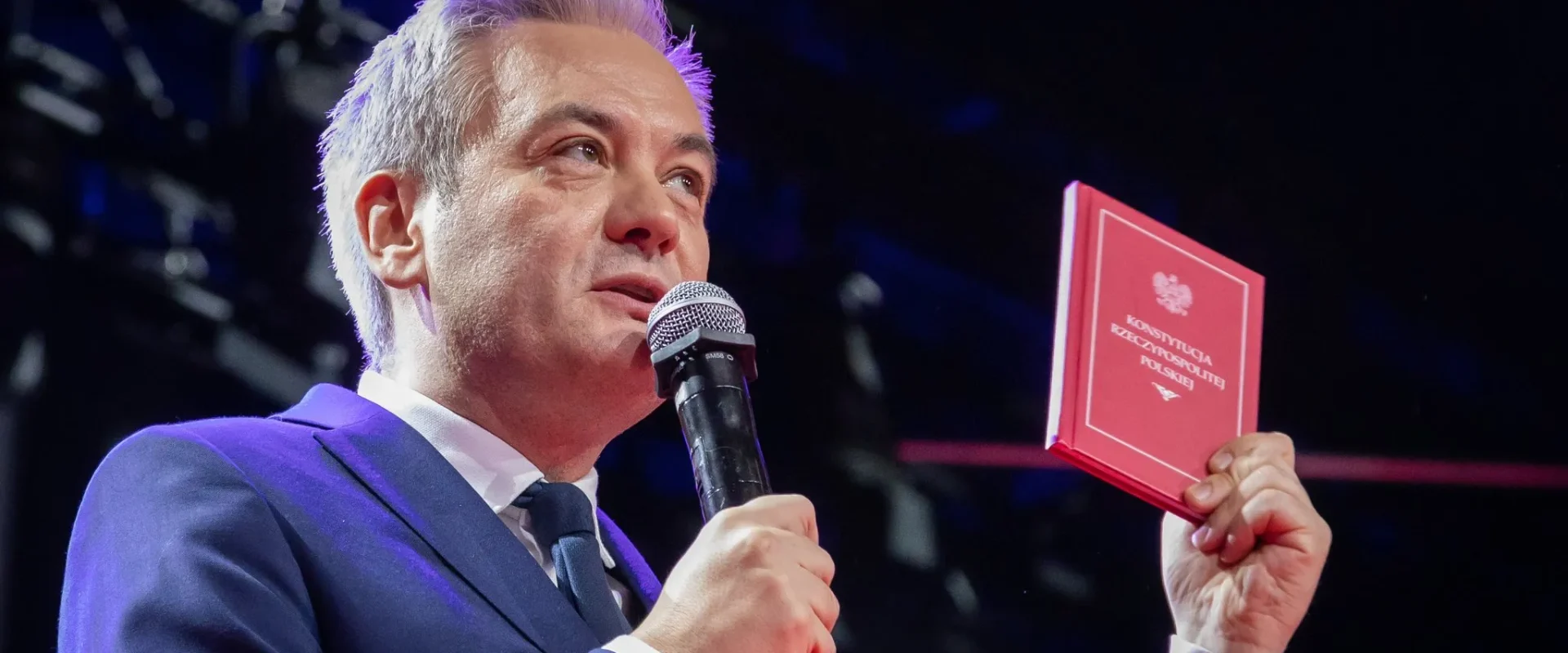 Mężczyzna w granatowym garniturze trzyma mikrofon w jednej ręce i czerwony podręcznik Konstytucji RP w drugiej, stojąc na scenie.