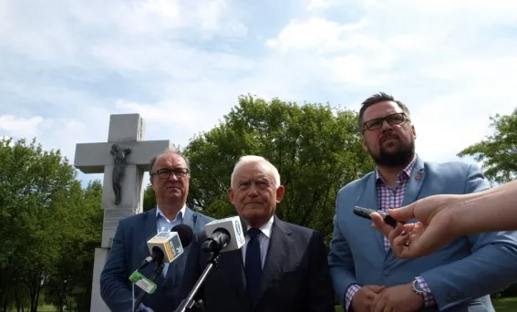 Trzech mężczyzn w garniturach stoi przy mikrofonach przed kamiennym krzyżem w parku, a z boku widać dłoń trzymającą dodatkowy mikrofon.