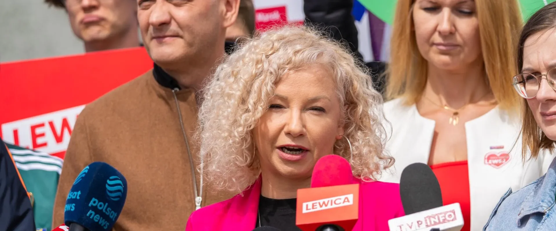 Kobieta o blond kręconych włosach w różowym żakiecie przemawia do mikrofonów podczas konferencji prasowej o ochronie konsumentów.