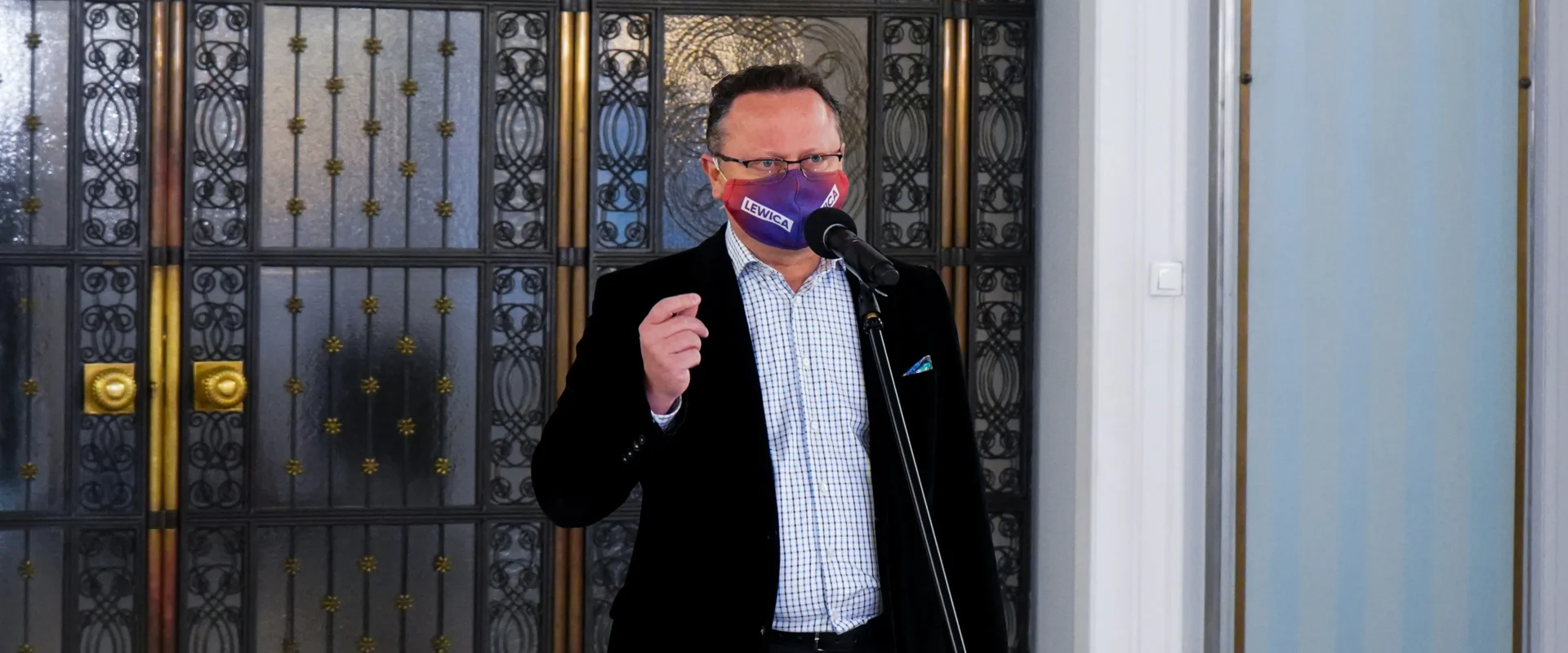 Mężczyzna w garniturze, z maseczką z napisem LEWICA, przemawia do mikrofonu w pomieszczeniu z ozdobnymi kutymi drzwiami w tle.