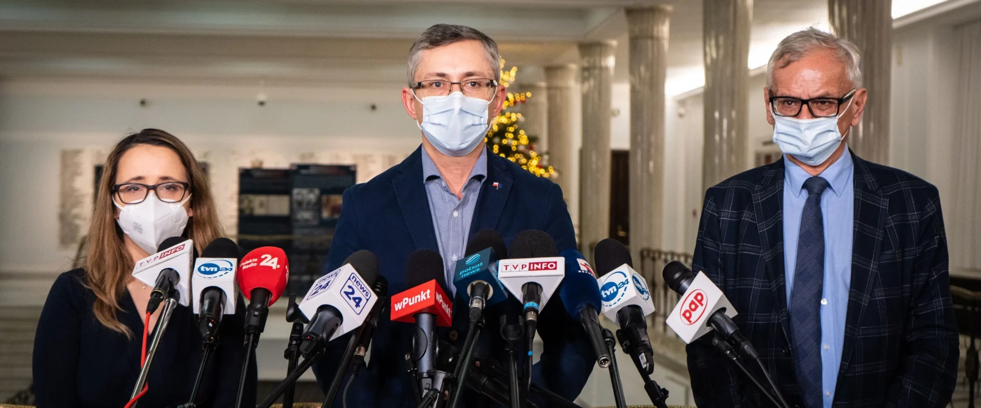 Trzech polityków w maseczkach stojących przed mikrofonami podczas konferencji prasowej w eleganckim wnętrzu.