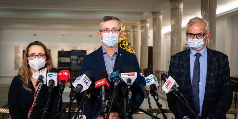 Trzech polityków w maseczkach stojących przed mikrofonami podczas konferencji prasowej w eleganckim wnętrzu.