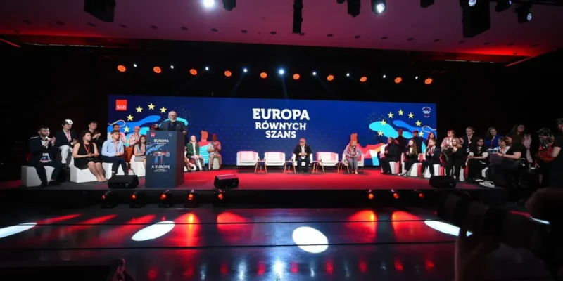 Zdjęcie przedstawia konferencję z hasłem "Europa Równych Szans", na której przemawia mężczyzna, a publiczność siedzi na białych kanapach w tle podświetlonym kolorowymi światłami.