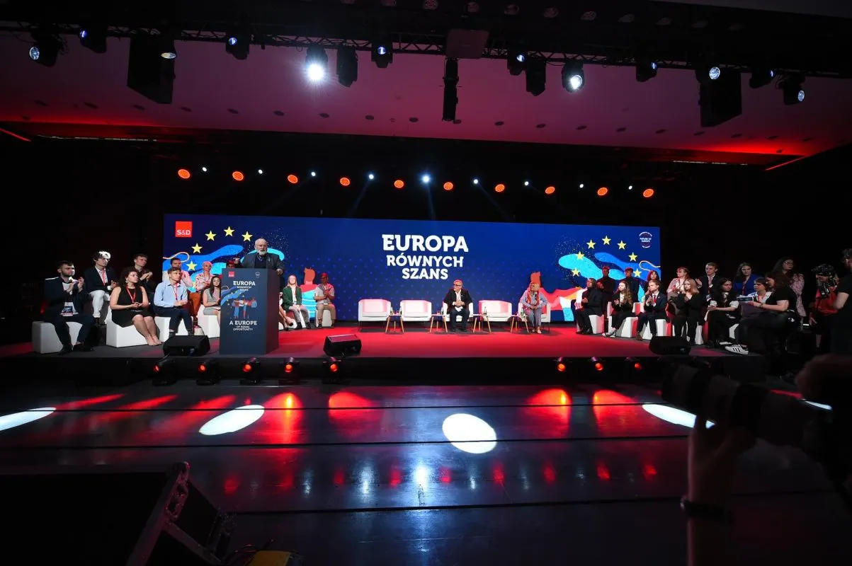 Zdjęcie przedstawia konferencję z hasłem "Europa Równych Szans", na której przemawia mężczyzna, a publiczność siedzi na białych kanapach w tle podświetlonym kolorowymi światłami.