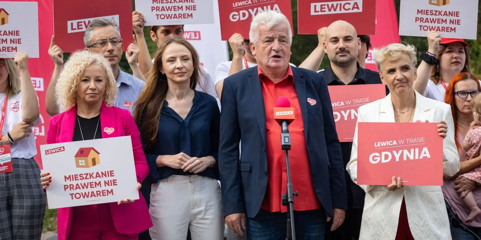 Grupa osób na wiecu Lewicy trzyma czerwono-białe plakaty z napisem LEWICA i hasłami, a mężczyzna w czerwonej koszuli i granatowej marynarce przemawia do mikrofonu.