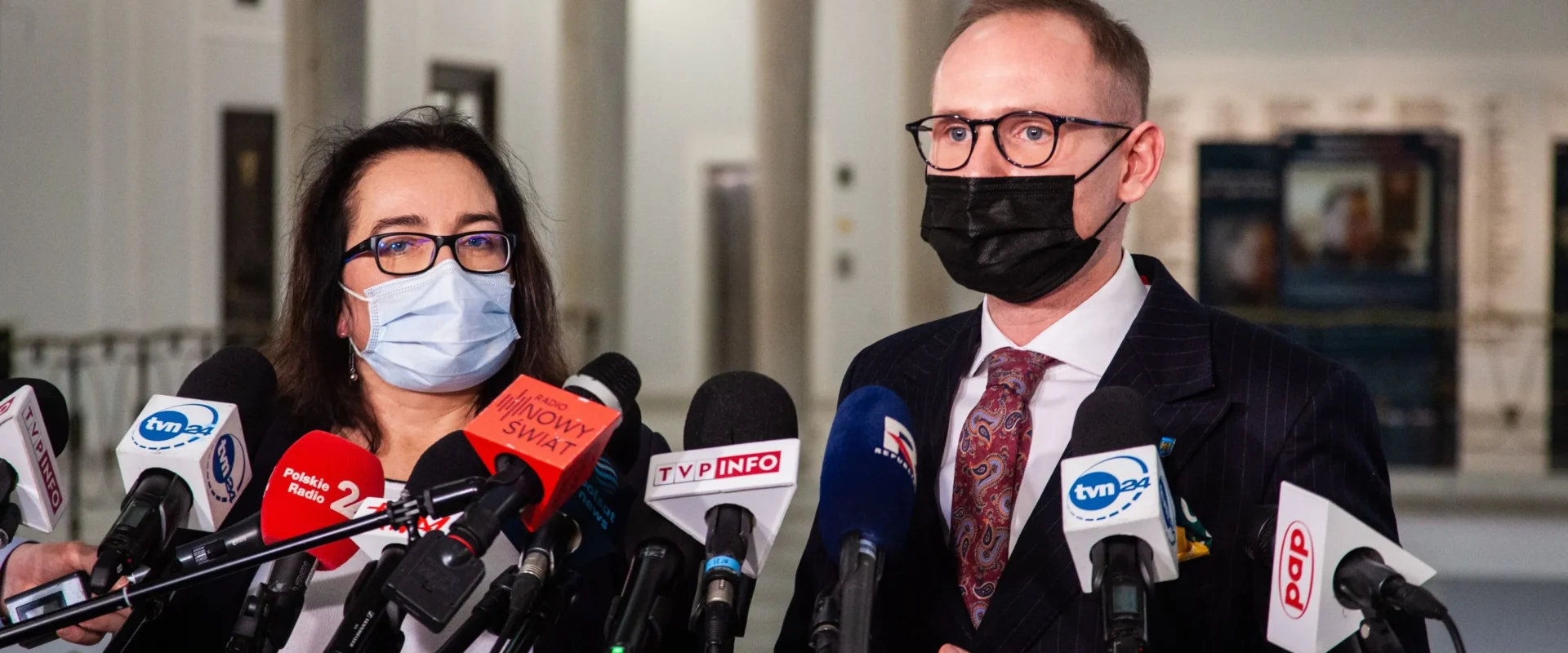 Dwóch urzędników w maseczkach udziela konferencji prasowej w Ministerstwie Sportu i Turystyki, stojąc przed licznymi mikrofonami mediów.