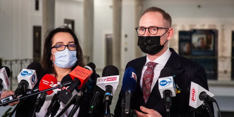 Dwóch urzędników w maseczkach udziela konferencji prasowej w Ministerstwie Sportu i Turystyki, stojąc przed licznymi mikrofonami mediów.