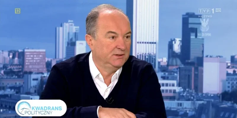 Mężczyzna w białej koszuli i ciemnym swetrze rozmawia w studiu telewizyjnym „Kwadrans Polityczny” na tle panoramy miasta.