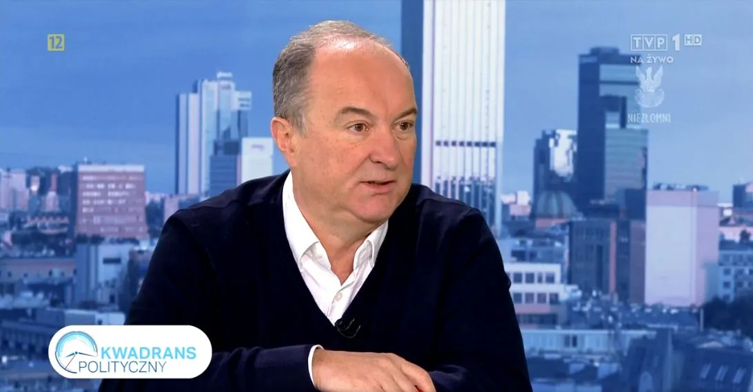 Mężczyzna w białej koszuli i ciemnym swetrze rozmawia w studiu telewizyjnym „Kwadrans Polityczny” na tle panoramy miasta.