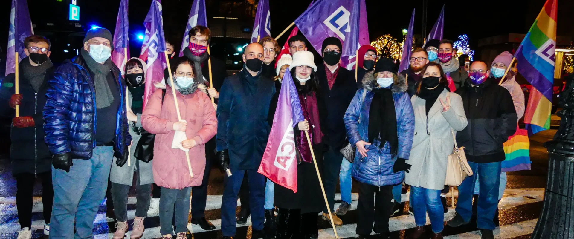 Grupa osób w zimowych ubraniach stoi na nocnym placu, trzymając fioletowe flagi i tęczową flagę podczas demonstracji przeciw Lex TVN.