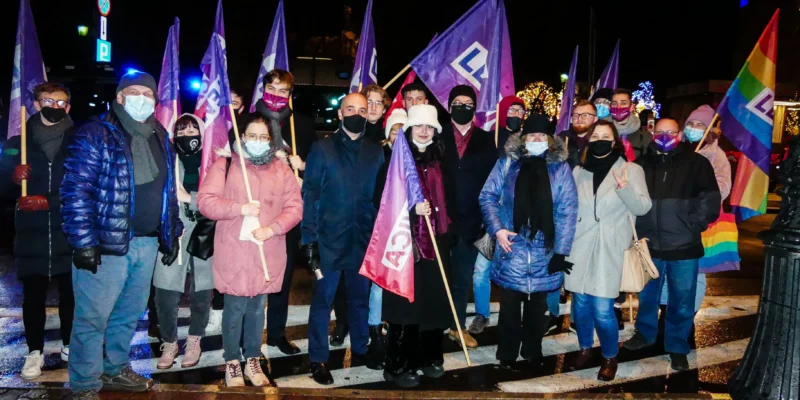 Grupa osób w zimowych ubraniach stoi na nocnym placu, trzymając fioletowe flagi i tęczową flagę podczas demonstracji przeciw Lex TVN.
