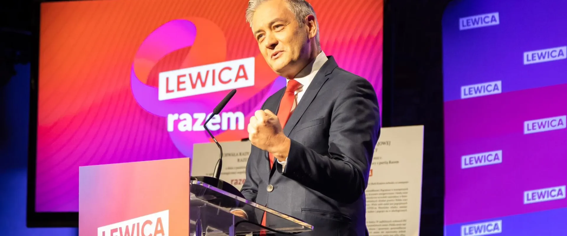 Na zdjęciu widać polityka przemawiającego z mównicy podczas wydarzenia związanego z partią Lewica, z logo „razem” w tle.