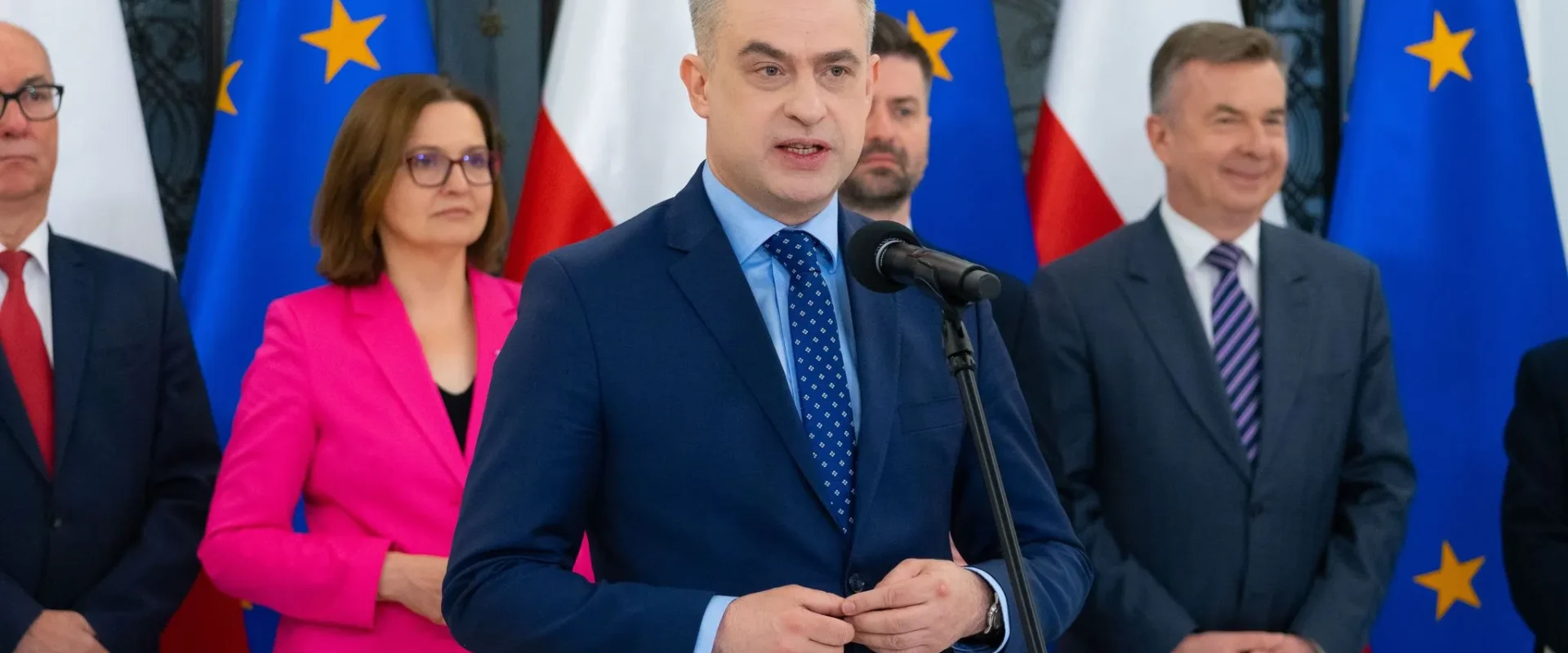 Polityk w granatowym garniturze przemawia do mikrofonu podczas konferencji prasowej, z flagami UE i Polski w tle oraz innymi urzędnikami stojącymi za nim.