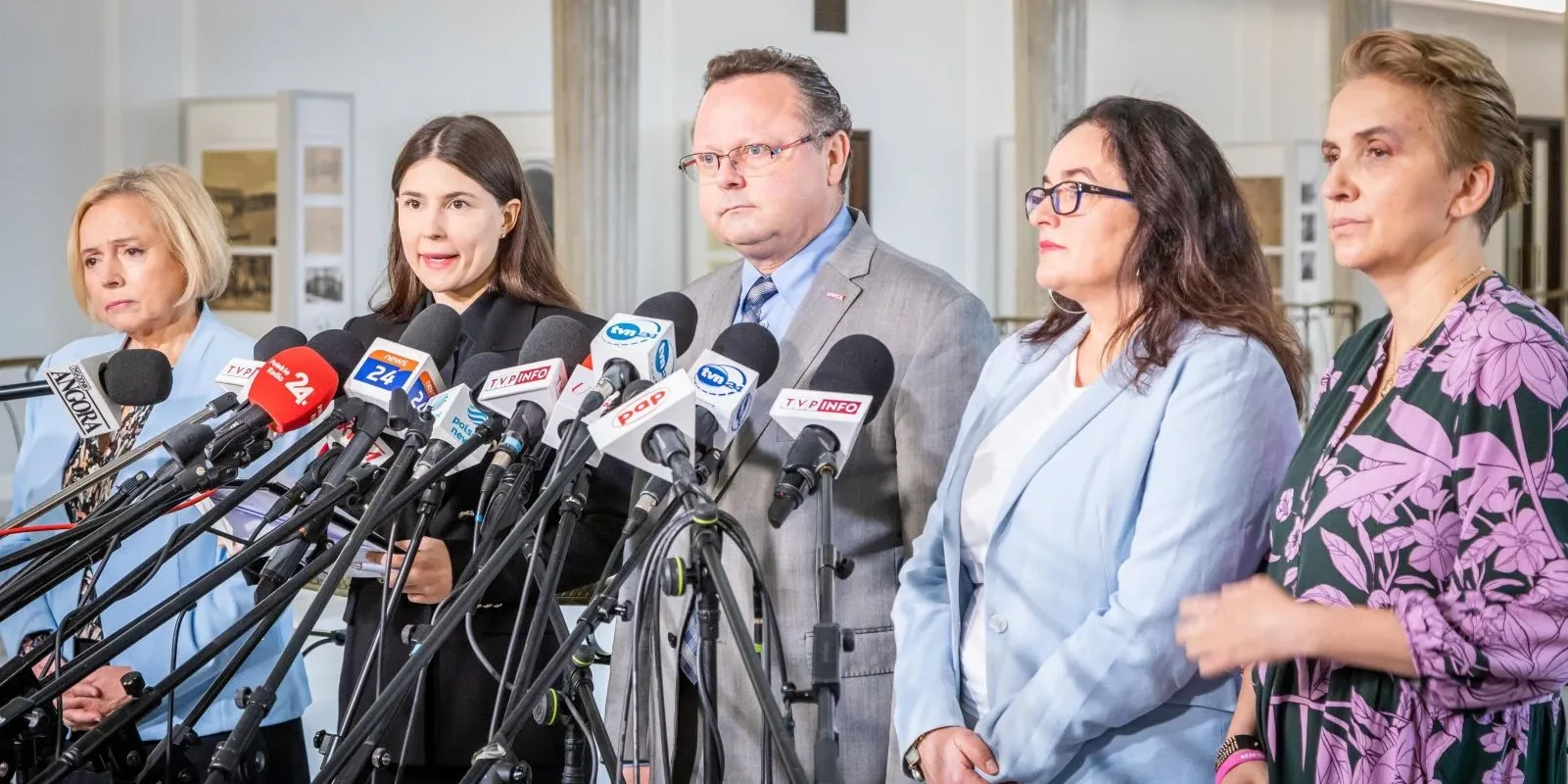 Na zdjęciu widoczna jest grupa pięciu osób stojących przy mikrofonach na konferencji prasowej.