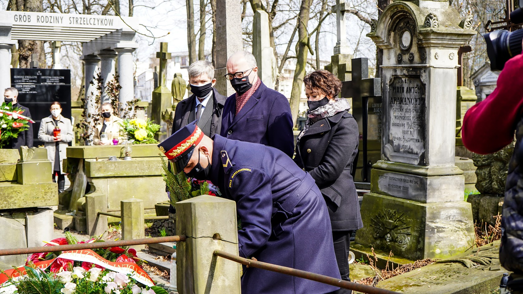 Grupa osób w maseczkach składa wieńce przy grobie na cmentarzu podczas obchodów 11. rocznicy katastrofy smoleńskiej.