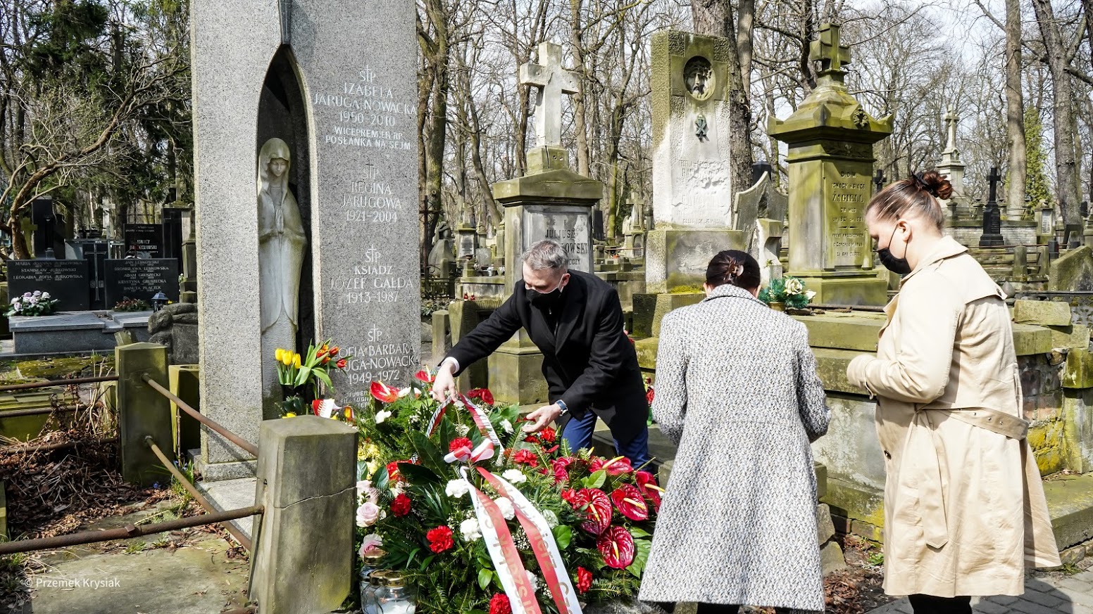 Grupa osób w maseczkach składa kwiaty przy grobie na cmentarzu, upamiętniając 11. rocznicę tragedii smoleńskiej.
