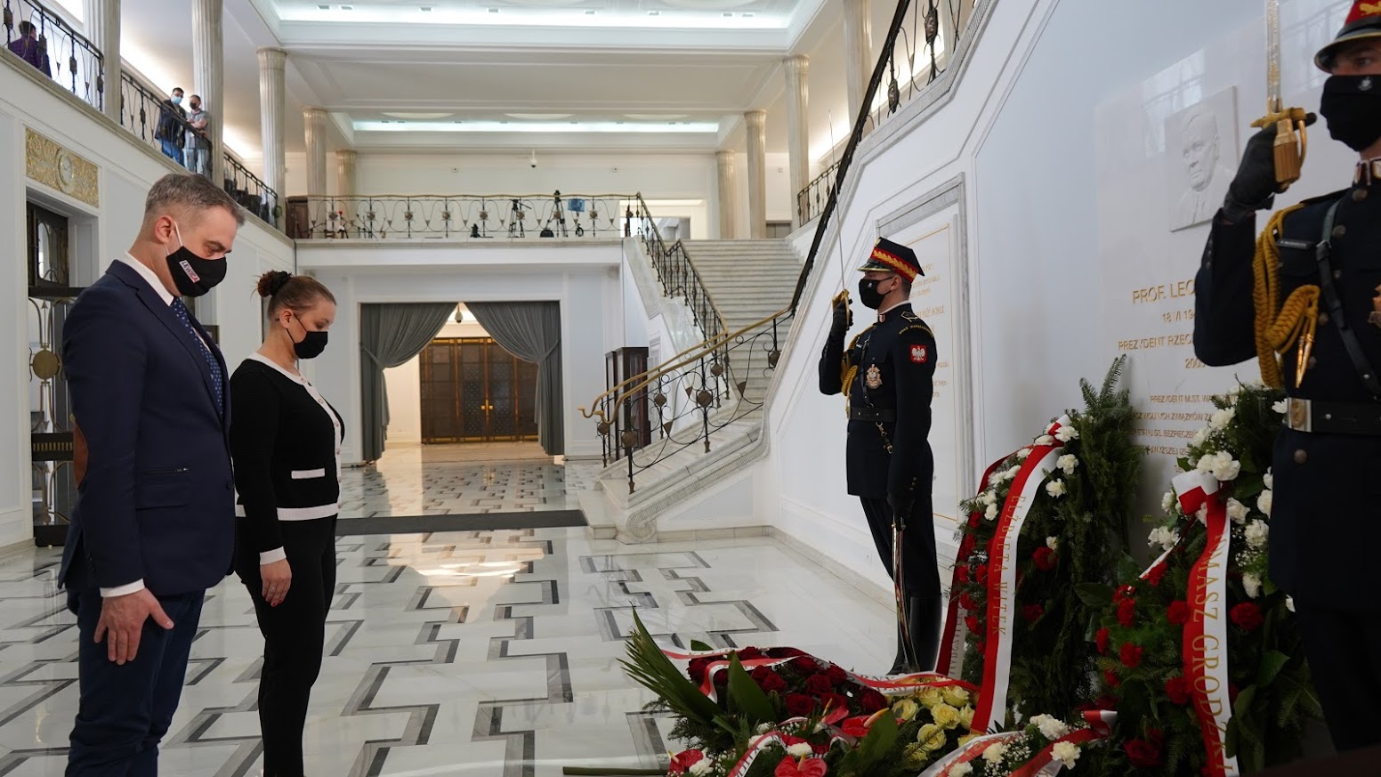 Para w maseczkach składa hołd w eleganckiej, marmurowej hali podczas obchodów 11. rocznicy tragedii smoleńskiej; po prawej stoją żołnierze w mundurach ceremonialnych przy wieńcach.