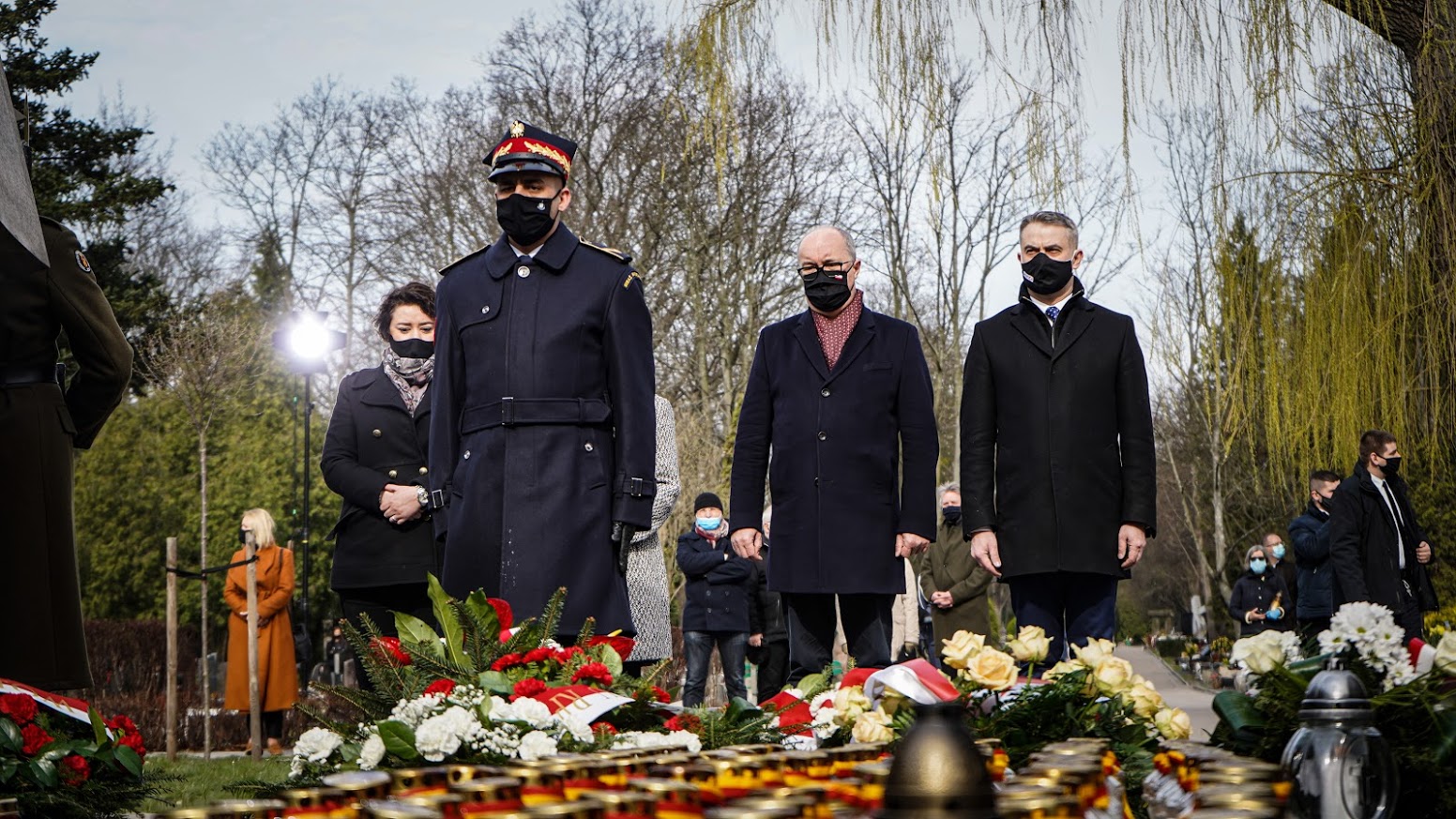 Grupa osób w maseczkach stoi przed kwiatami i zniczami podczas uroczystości upamiętniającej 11. rocznicę tragedii smoleńskiej.