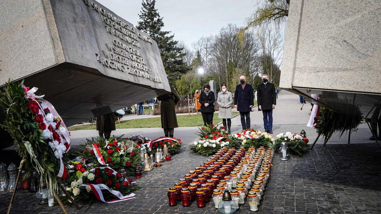 Uroczystość upamiętniająca 11. rocznicę tragedii smoleńskiej – kilka osób w maskach stoi przed monumentalnym pomnikiem, otoczonym kwiatami i zniczami.