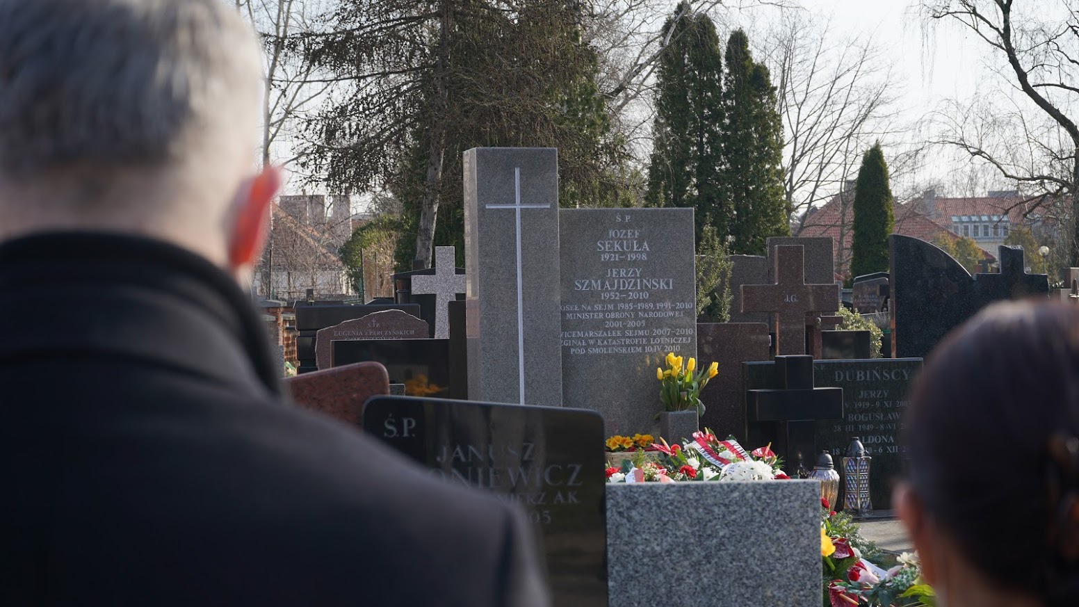 Dwie osoby stoją na cmentarzu przy grobach z kwiatami i krzyżami, upamiętniając 11. rocznicę tragedii smoleńskiej.