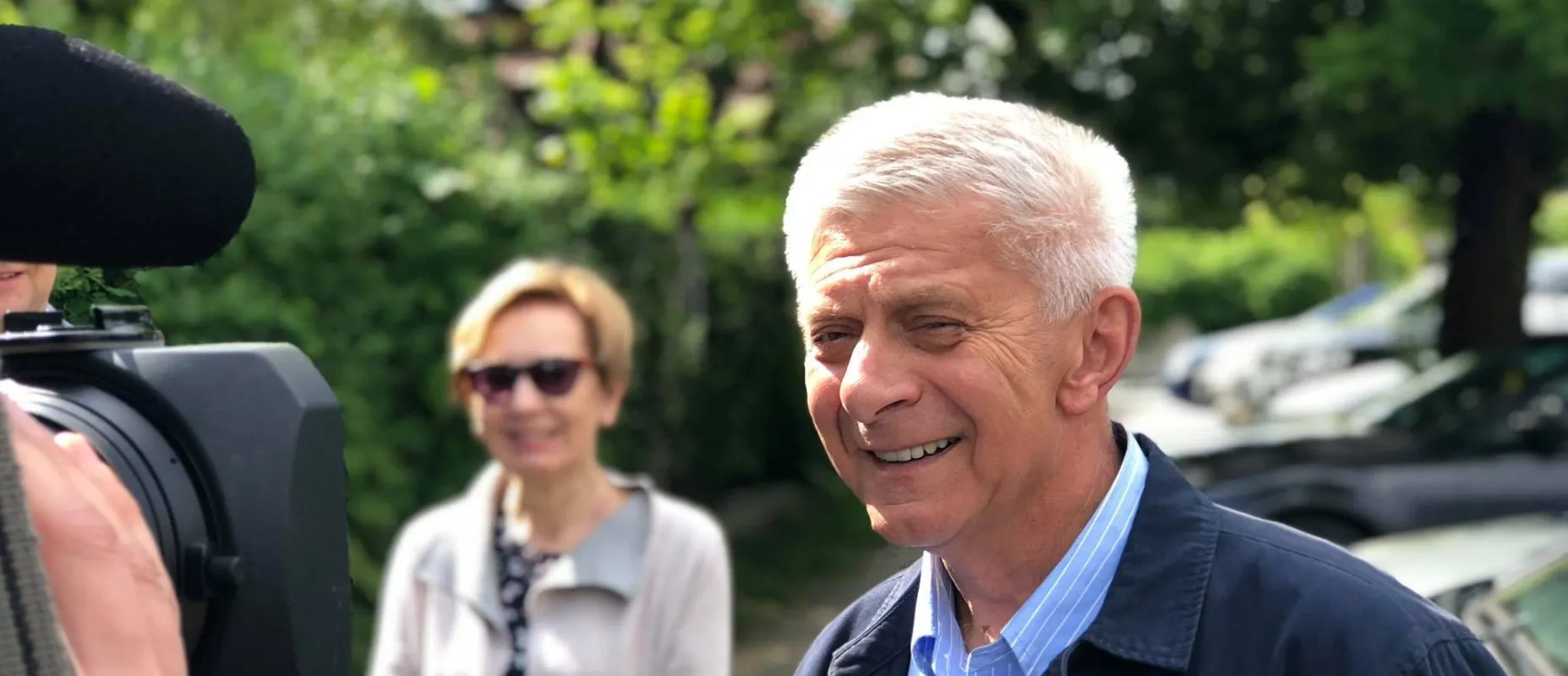 Uśmiechnięty starszy mężczyzna o siwych włosach stoi na zewnątrz obok kamery, a w tle widać kobietę w okularach.