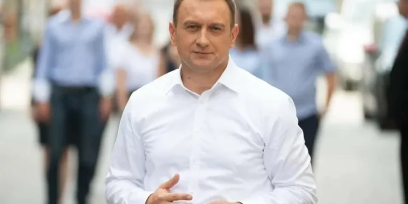 Mężczyzna w białej koszuli stoi na ulicy, a w tle widoczni są przechodnie.