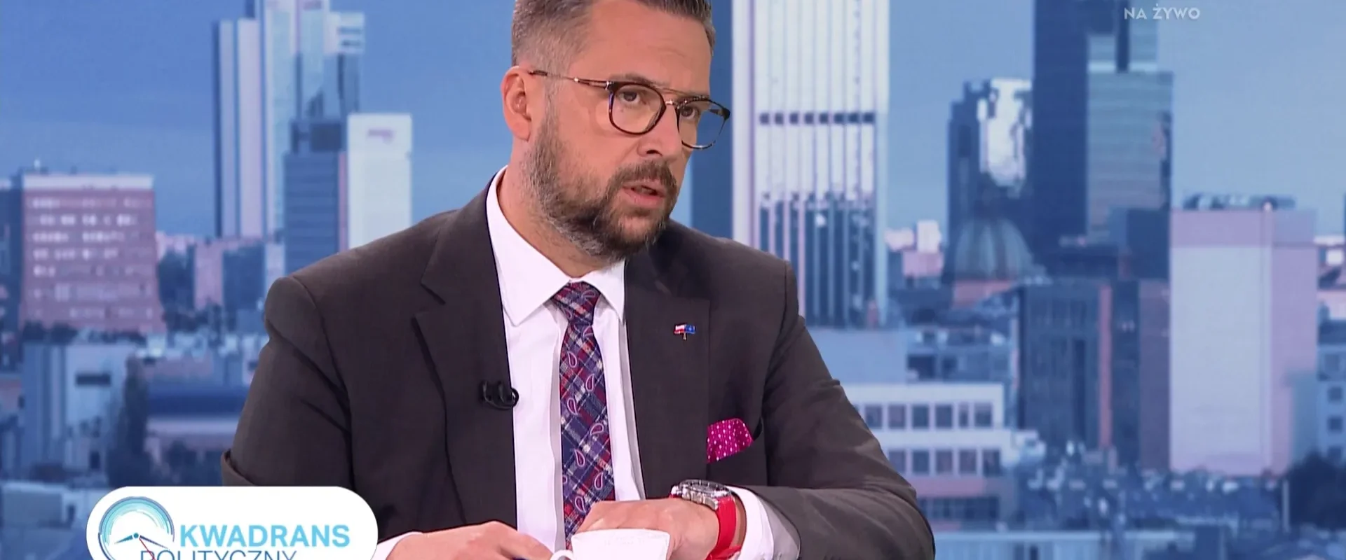 Mężczyzna w garniturze siedzi przy stole z filiżanką kawy, prowadząc rozmowę na tle nowoczesnej miejskiej panoramy.