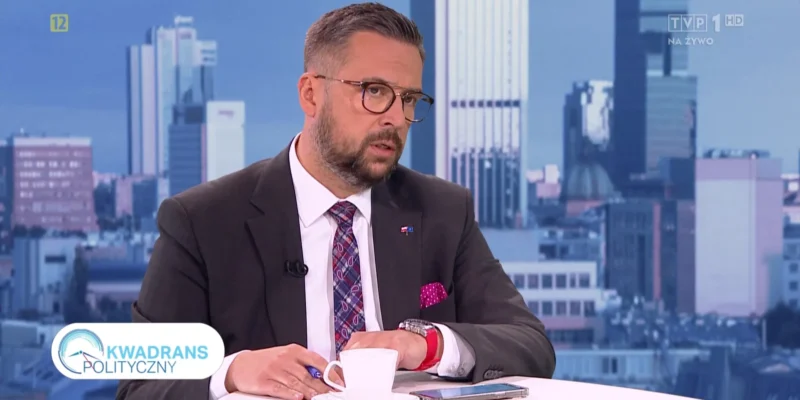 Mężczyzna w garniturze siedzi przy stole z filiżanką kawy, prowadząc rozmowę na tle nowoczesnej miejskiej panoramy.