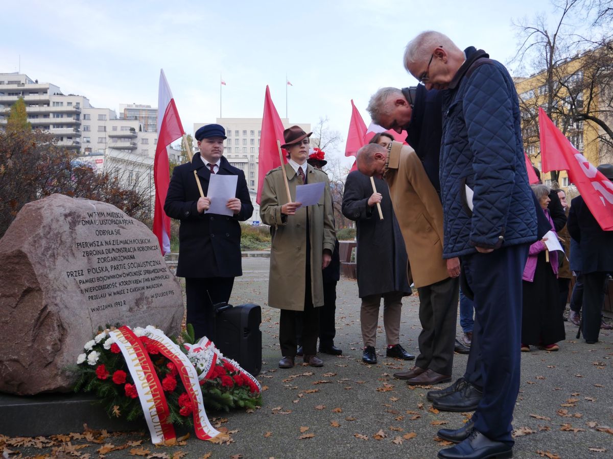 Grupa osób bierze udział w obchodach Narodowego Święta Niepodległości, stojąc przy kamiennym pomniku z wieńcami i czerwonymi flagami, odczytując tekst z kartek.