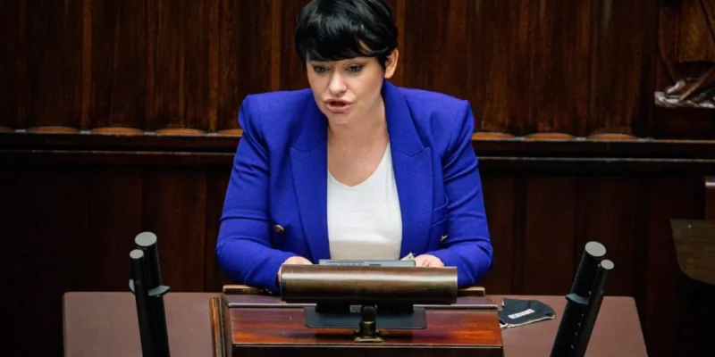 Kobieta w niebieskim żakiecie przemawia z mównicy w sali obrad.