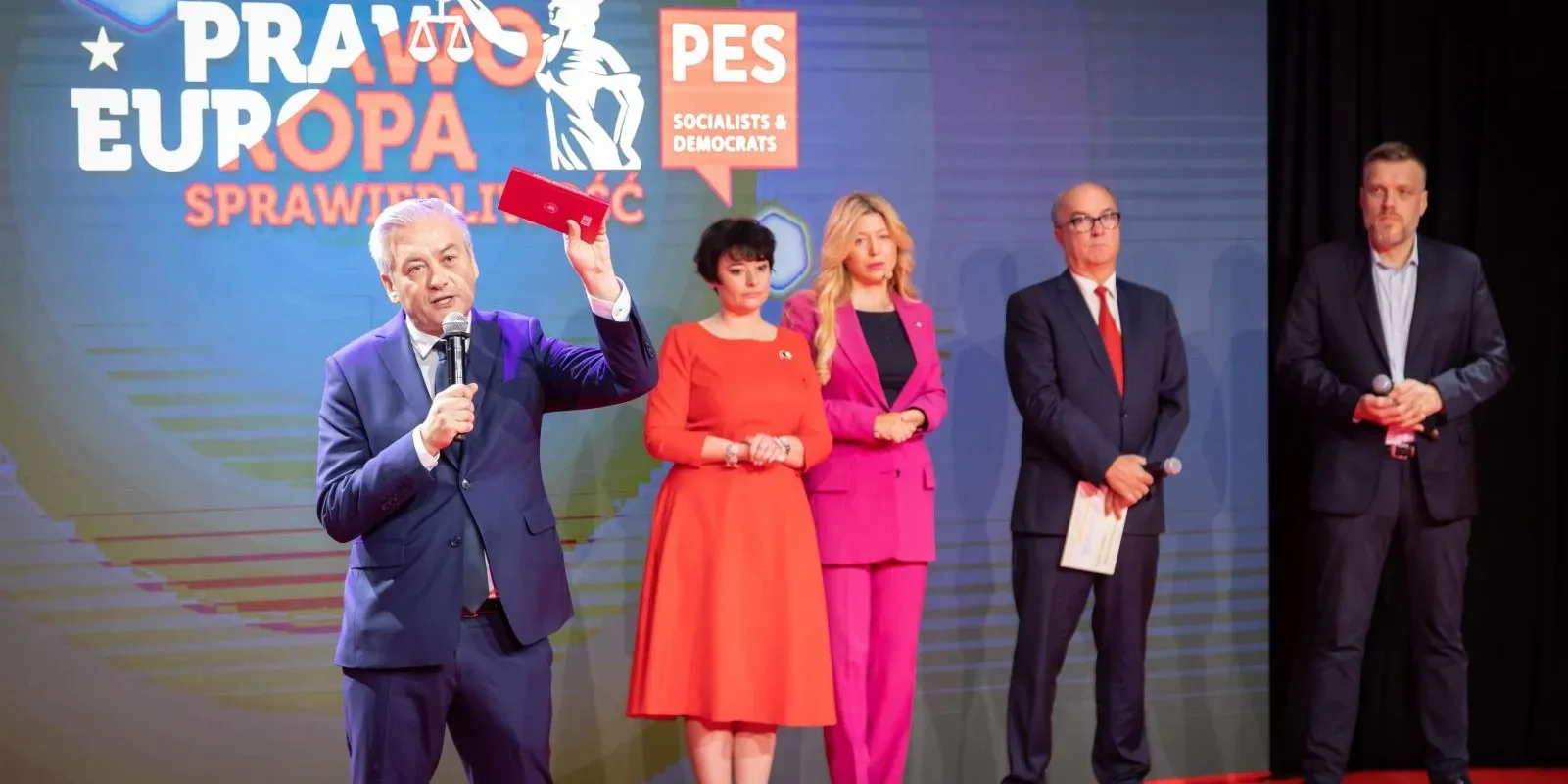 Na zdjęciu widoczny jest mężczyzna przemawiający do mikrofonu na tle tła z logo konwencji, obok niego stoi grupa osób w różnych kolorowych strojach.