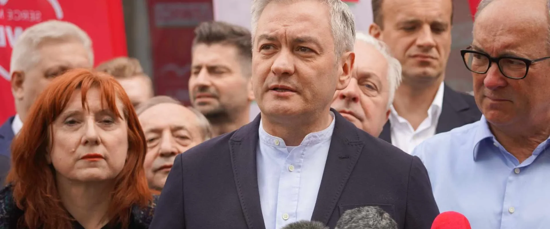 Mężczyzna w granatowej marynarce przemawia do mediów podczas konferencji prasowej, otoczony innymi osobami i dziennikarzami, z mikrofonami przed nim i czerwonymi banerami w tle.