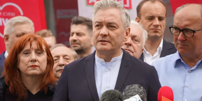 Mężczyzna w granatowej marynarce przemawia do mediów podczas konferencji prasowej, otoczony innymi osobami i dziennikarzami, z mikrofonami przed nim i czerwonymi banerami w tle.