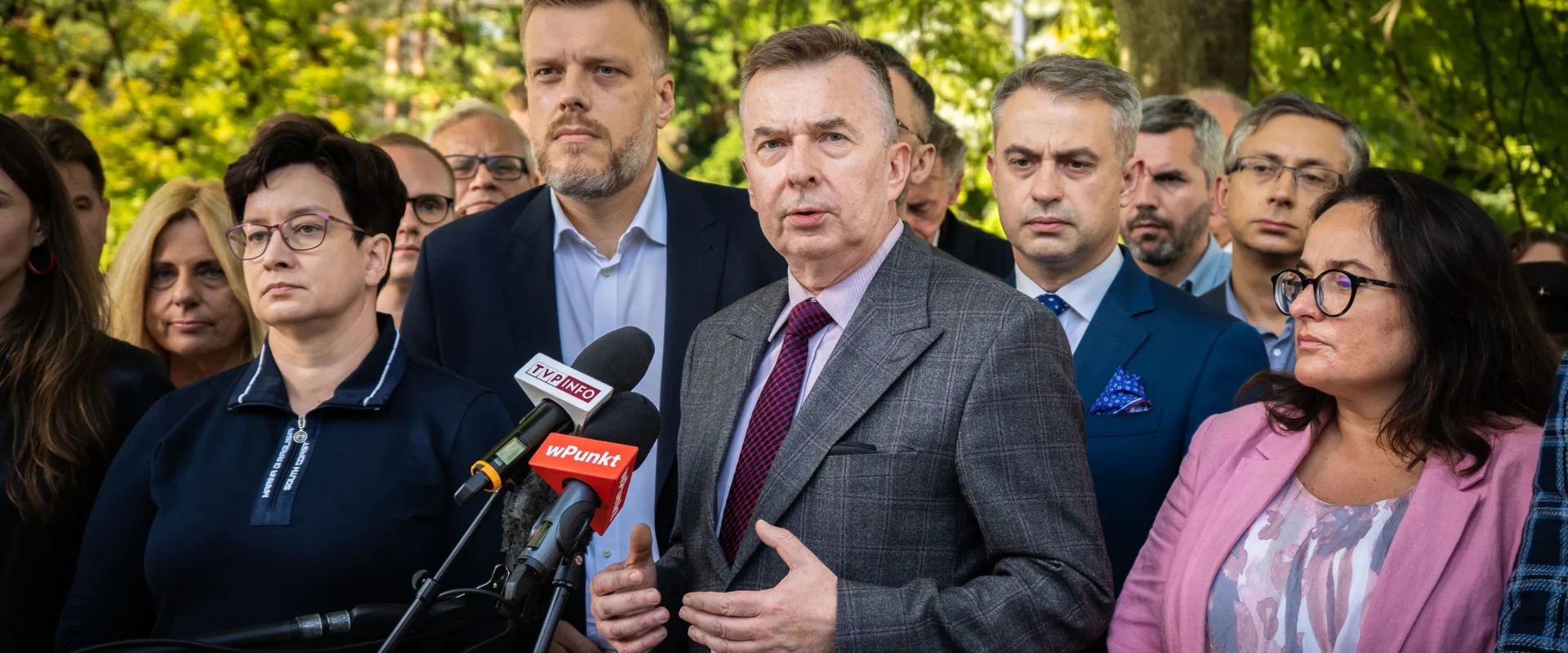 Grupa polityków na zewnątrz w parku podczas konferencji prasowej, centralnie przemawia mężczyzna w kraciastym garniturze i fioletowym krawacie, otoczony mikrofonami z logotypami TVP Info i wPunkt.