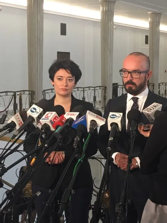 Dwie osoby w formalnych strojach udzielają konferencji prasowej przed wieloma mikrofonami w reprezentacyjnym wnętrzu, komentując sprawy legislacyjne.